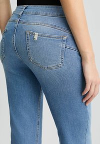 Jeans en denim bleu clair avec une taille mi-haute, dotés de poches arrière et de détails de couture subtils. Le tissu a une texture lisse.