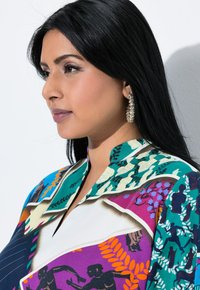 Bunte gemusterte Bluse mit einer Mischung aus lila, grün und blauen Designs, klassischem Kragen und markanten floralen sowie menschlichen Motiven.