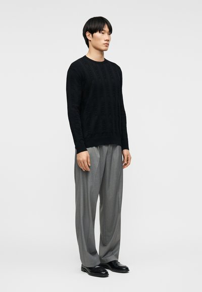 Jeune homme debout, portant un pull noir texturé à manches longues, un pantalon gris ample et des chaussures habillées noires, sur un fond clair uni.