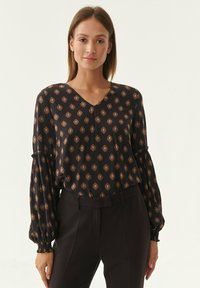 Schwarze Bluse mit V-Ausschnitt, die ein durchgehendes Diamantmuster in Braun aufweist. Lange Ärmel mit gerafften Manschetten und leichtem Stoff.