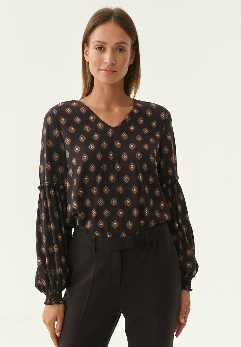 Schwarze Bluse mit V-Ausschnitt, die ein durchgehendes Diamantmuster in Braun aufweist. Lange Ärmel mit gerafften Manschetten und leichtem Stoff.