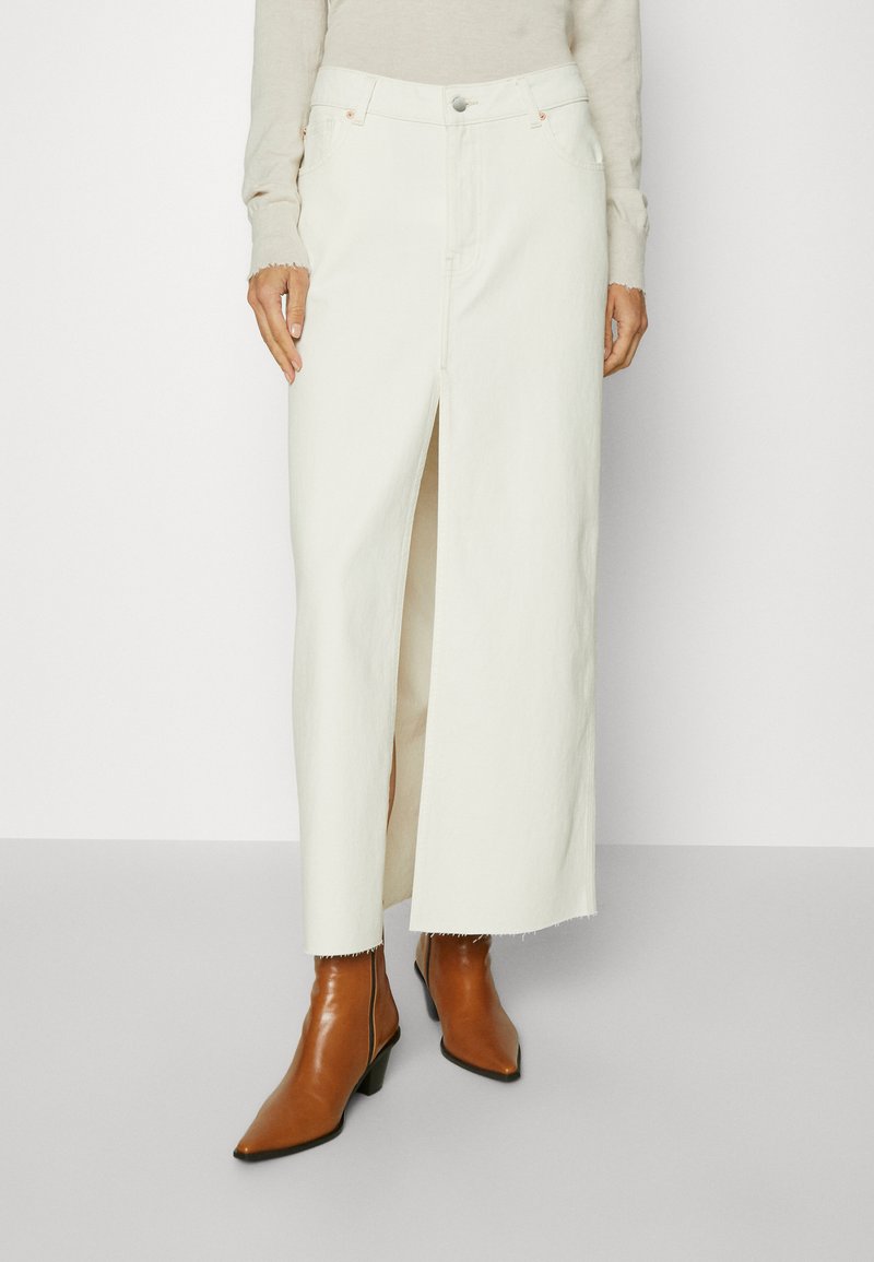 Witte denim rok met een voorzijde split, hoge taille en franjes aan de onderkant; gecombineerd met bruine enkellaarzen met een spitse neus en blokhak.