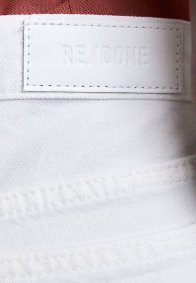 Tela de denim blanco con una superficie texturizada, presenta un parche de cuero etiquetado "RE/DONE" cosido en la cinturilla.