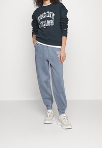 Sudadera azul oscura con gráfico de "TOMMY JEANS", combinada con joggers gris claros con impresión de logo y zapatillas beige con acentos grises.