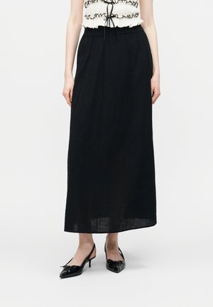 Γυναίκα που φορά μαύρη φούστα μήκους μέχρι τον αστράγαλο, λευκό αμάνικο τοπ με βολάν και μαύρο περίβλημα, και μαύρα γόβες slingback με μύτη και φιόγκους.