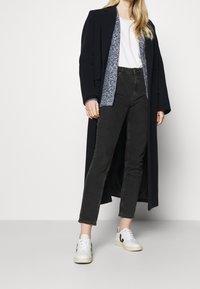 Lång marinblå kappa över en grå stickad cardigan, vit t-skjorta, svarta jeans och vita sneakers med svarta detaljer. Platta skor och avslappnad passform.
