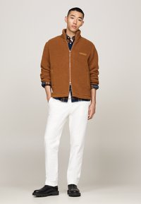 Brun fleece jakke med glidelås og sidelommer, brukt over en blå rutete skjorte og hvite bukser, kombinert med svarte støvler med kraftig såle.