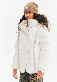 Biała pikowana kurtka puchowa z przeszywanym wzorem, beżowymi akcentami, wysokim kołnierzem i zamkiem błyskawicznym, w połączeniu z brązową dzianinową czapką.