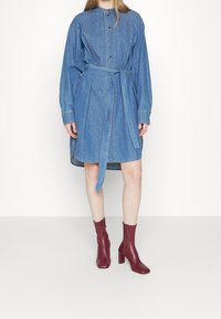 Denim shirt dress with a button-up front, long sleeves, and a tie belt at the waist, featuring a curved hem and blue color. Paired with burgundy ankle boots.