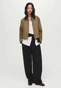 Veste en coton marron avec un col, associée à une chemise blanche à rayures et un pantalon large noir. Des chaussures noires complètent la tenue.