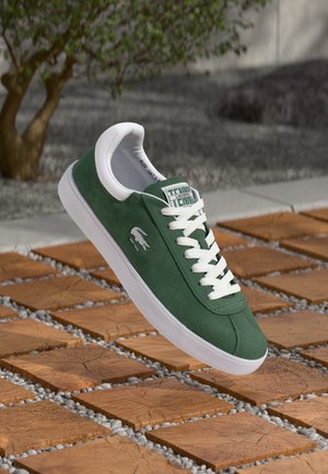 Zelený semišový teniska s bílou podrážkou a tkaničkami, logo Lacoste na boku, zobrazená nad dřevěnou dlažbou a štěrkovým venkovním povrchem.
