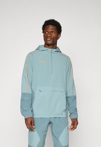 Light blue windbreaker with a half-zip design, orange accent seams, front pocket, and adjustable hood. Made from lightweight fabric.
