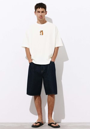Ein junger Mann trägt ein weißes T-Shirt mit Hamster-Grafik, dunkle Jeansshorts und schwarze Flip-Flops, steht an einer weißen Wand mit den Händen in den Taschen.