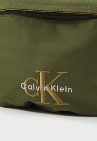 Borsa in tessuto verde oliva con un vistoso logo dorato "CK" e il testo "Calvin Klein" in bianco. Cerniera con dettagli di cucitura gialla.