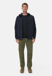 Marineblauer Hoodie mit Frontreißverschluss und Taschen, kombiniert mit olivfarbenen Cargo-Hosen mit Seitentaschen und braunen Sneakern mit einer strukturierten Oberfläche.