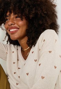 Femme souriante avec des cheveux bouclés portant un chandail beige avec de petits cœurs en paillettes brunes et des colliers dorés à couches.