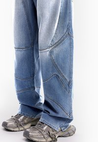 Svijetloplave traperice s jedinstvenim dizajnom prošivenih panela. Nosene s sivim sportskim cipelama, koje prikazuju teksturirani materijal i oštrih oblika.