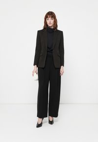 Mustat räätälöidyt bleiseri tumman turtleneckin päällä, leveälahkeiset mustat housut ja mustat korot. Pitelee pientä valkoista laukkua. Yksinkertainen muotoilu, rakenteellinen istuvuus.
