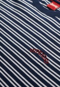 Tissu à rayures bleu marine et blanc avec texte brodé rouge "Superdry" et une étiquette de marque rouge visible près du col.