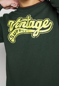 Zelená mikina s dlouhým rukávem, která má na hrudi výrazný vybledlý žlutý grafický potisk "Vintage Supply" v tučném písmu. Měkká textura.