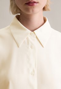 Κρέμα χρώματος πουκάμισο με κουμπιά, φτιαγμένο από λείο ύφασμα, με κλασικό γιακά και δύο ορατά κουμπιά μπροστά.