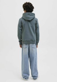 Sudadera verde con capucha grande en la parte de atrás, acompañada de jeans anchos de color azul claro y zapatillas blancas. Textura suave con costuras visibles y bolsillos.