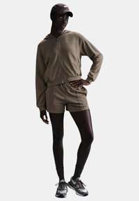 Felpa khaki con zip e pantaloni corti abbinati, realizzati in tessuto morbido, dal fitting rilassato. Abbinati a sneaker bianche e marroni e a un cappellino marrone.