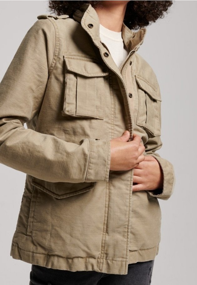 Personne aux cheveux bouclés portant une veste utilitaire beige avec des poches à rabat, fermant la fermeture éclair avant sur une chemise blanche et un pantalon foncé.