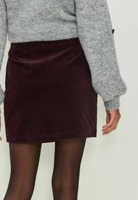 Bordová kordurová minisukňa s hladkou textúrou a jednoduchým dizajnom, skombinovaná so svetlošedým pleteným sveterom a čiernymi pančuchami.