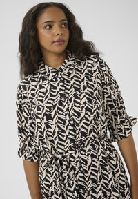 Black and beige shirt dress with a zigzag pattern, short ruffled sleeves, button-down front, and a tie belt at the waist.