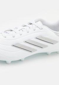 Baltas futbola apavi ar teksturētu sintētisko virsmu, trim metāliskām sudraba svītrām un auklu aizdari ar veidotiem spike.