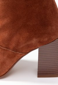 Bottine en suède marron avec un talon haut effilé. Le talon présente une texture ressemblant à du bois avec des rainures horizontales. Détail de couture visible sur le dessus.