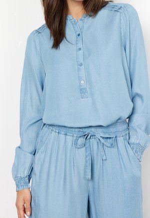 Femme portant une chemise bleu clair à manches longues boutonnée et un pantalon assorti à cordon, poignets et épaules smockés, les mains détendues le long du corps.