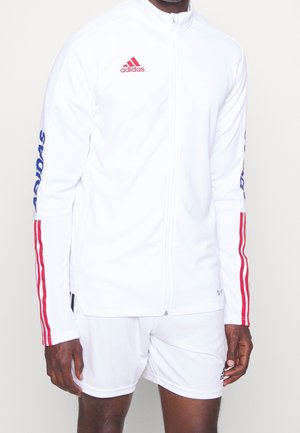 Mand iført hvid Adidas træningsjakke med røde striber på ærmerne og matchende hvide shorts, stående mod en ensfarvet baggrund.