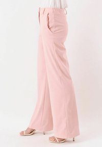 Persona che indossa pantaloni rosa pallido a vita alta e gamba larga con tasche laterali e sandali beige con tacco, in piedi davanti a uno sfondo bianco.