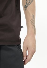 Hnědé bavlněné tričko s krátkými rukávy, které ukazuje lem a černou etiketu na boku; kombinováno s černými kalhotami a tetovanou rukou.