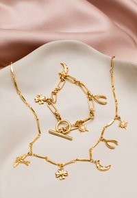 Conjunto de joyería en tono dorado que incluye un collar y una pulsera con charms como lunas, estrellas, un pájaro y un trébol de cuatro hojas. Textura suave, diseño entrelazado.