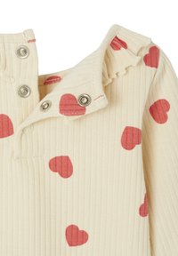 Rippiges cremefarbenes Kleidungsstück mit roten Herzmustern, einem Knopfkragen und Rüsche entlang des Ausschnitts. Weicher Stoff, lässiges Design.