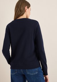 Sweatshirt bleu marine à manches longues, avec des poignets et un ourlet côtelés. Tissu doux, col rond, et design uni au dos. Associé à un jean bleu.