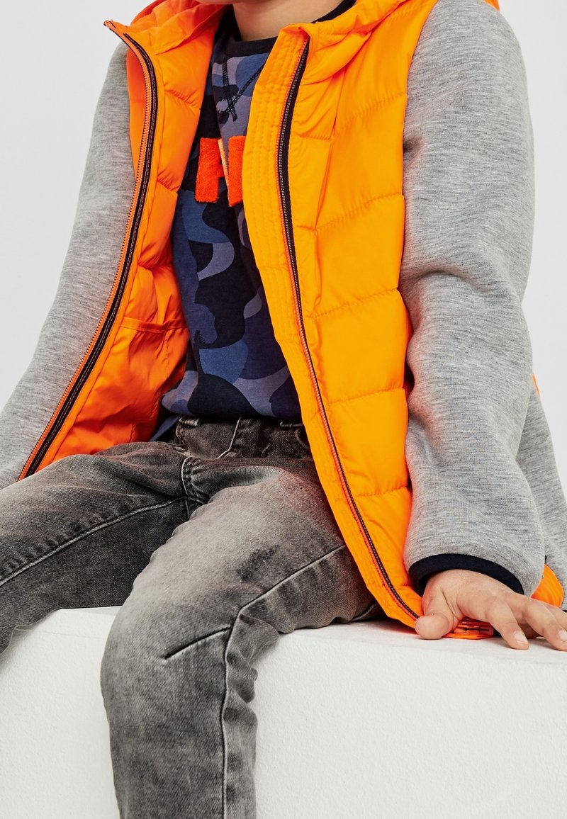 Enfant portant un gilet orange par-dessus un sweat à capuche gris et un jean foncé, assis avec la main posée sur une surface blanche.