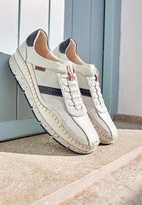 Weiße Freizeitsneaker mit navyblauen Streifen, beigen Nähten und dicken gesprenkelten Sohlen auf einer Steinoberfläche neben einer blauen Holztür.