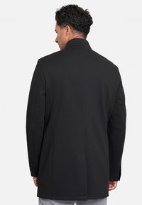 Abrigo negro con un cuello estructurado, ajuste entallado y puños de tres botones. Textura suave y diseño minimalista. Presenta costuras pronunciadas.