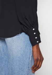 Schwarzes Langarmshirt mit gerafften Ärmeln, ausgestattet mit dekorativen Knöpfen und einer weichen Textur, kombiniert mit hellblauen Jeans.