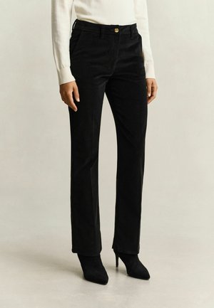 Femme portant un pantalon noir droit, un haut blanc à manches longues et des bottines noires à talons, debout sur un sol et un fond clairs et unis.