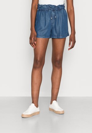 Personne portant un short en jean bleu avec une taille élastique et des baskets blanches sur fond uni.