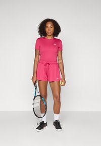 Camisola atlética rosa de mangas curtas, calções com cordão à medida, sapatilhas pretas, segurando uma raquete de ténis e uma bola. Acabamentos com logótipos brancos.