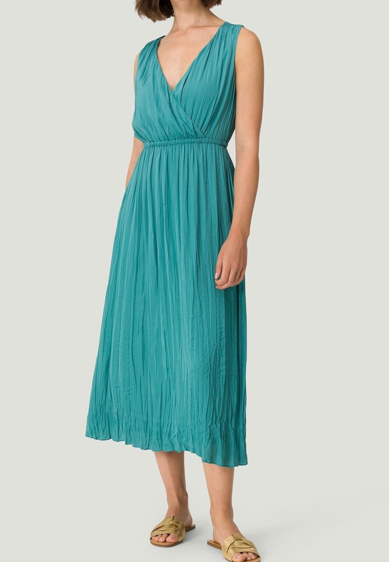 Teal ærmeløs omslagskjole med v-udskæring, plisseret design og elastisk talje; stoffet draperer fra taljen til anklerne, parret med gyldne sandaler.