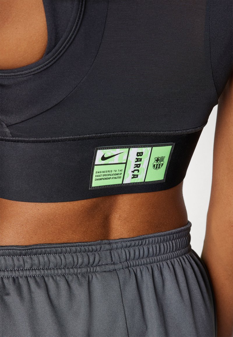 Primo piano di una persona che indossa un reggiseno sportivo nero con etichette verdi Nike e Barça, e pantaloni corti grigi con fascia elasticizzata.