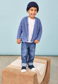 Cardigan tricotat albastru, tricou alb, blugi denim cu manșete răsucite și pantofi slip-on bleumarin. Copil zâmbitor stând pe blocuri texturate.