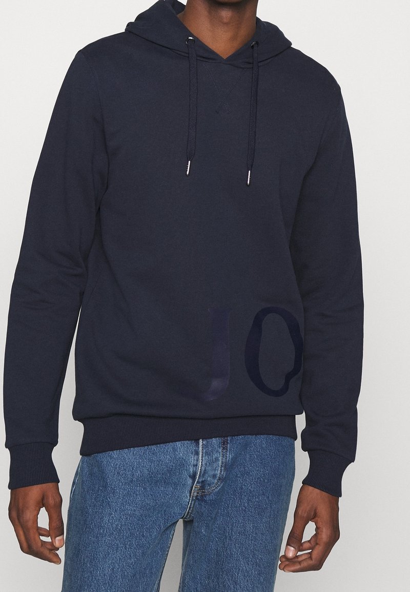 Man i en mörk marinblå hoodie med dragsnören och svaga "JO"-bokstäver, i kombination med blå denimjeans, stående mot en enkel bakgrund.